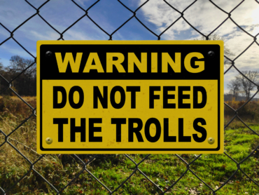 Black and yellow warning sign on a fence stating in "Warning - Do not feed the trolls".